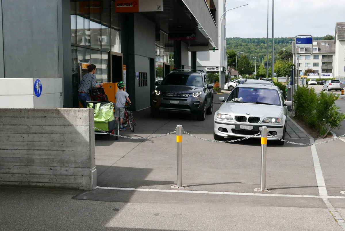Autos vor einer Postfiliale sowie eine Frau und ein Kind mit Velo.